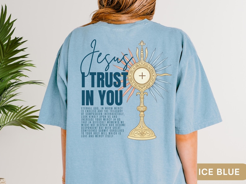Peut inclure: T-shirt bleu clair avec l'inscription "Jesus I Trust In You". Une monstrance dor&eacute;e avec une croix est repr&eacute;sent&eacute;e au dos, accompagn&eacute;e d'une citation religieuse. Le texte "ICE BLUE" est en bas.