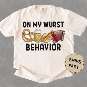 Può includere: Una t-shirt a maniche corte color crema con la scritta nera ON MY WURST BEHAVIOR. La grafica include un pretzel marrone, un boccale di birra, un hot dog nel panino e una fisarmonica rossa. La maglietta ha un girocollo e una vestibilità ampia.