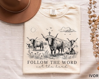 May include: Ivory-colored t-shirt featuring a black and white illustration of longhorn cattle in a field, with a cross and the sun in the background. The text reads "FOLLOW THE WORD not the herd."