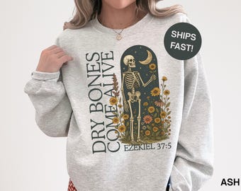 Pode incluir: Camisola cinza-claro com uma ilustração de esqueleto dentro de um arco, rodeada por girassóis e uma lua crescente. O texto "DRY BONES COME ALIVE" está impresso verticalmente à esquerda, com "EZEKIEL 37:5" por baixo.