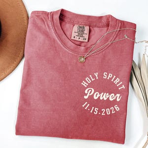 May include: A dusty rose Comfort Colors t-shirt with the words "HOLY SPIRIT POWER 11.15.2026" printed in a circular design. A gold pendant necklace and a brown hat are also visible.
