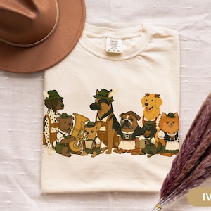 May include: Ivory-colored t-shirt featuring a whimsical illustration of various dog breeds dressed in traditional Bavarian attire. The dogs are depicted with hats, lederhosen, and musical instruments, creating a festive and playful design. A brown hat and dried flowers are in the background.