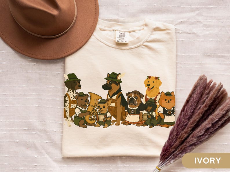 May include: Ivory-colored t-shirt featuring a whimsical illustration of various dog breeds dressed in traditional Bavarian attire. The dogs are depicted with hats, lederhosen, and musical instruments, creating a festive and playful design. A brown hat and dried flowers are in the background.
