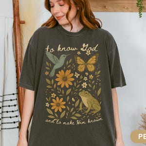 May include: Pepper-colored t-shirt featuring a nature-themed graphic. The design includes a hummingbird, butterfly, flowers, and a frog, with the text "to know God and to make Him known" in a vintage style.