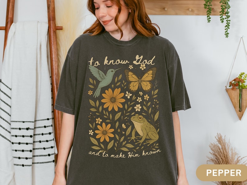 May include: Pepper-colored t-shirt featuring a nature-themed graphic. The design includes a hummingbird, butterfly, flowers, and a frog, with the text "to know God and to make Him known" in a vintage style.