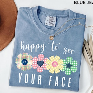 Puede incluir: Camiseta azul vaquero con la frase "happy to see your face" en blanco. El diseño incluye cuatro motivos florales con detalles de botones y corazones. Un collar dorado está sobre la camiseta.