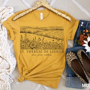 Pode incluir: Camiseta amarelo mostarda com um gr&aacute;fico de paisagem preto e o texto "ST. THERESE OF LISIEUX ora pro nobis." A camisa &eacute; combinada com jeans, sapatilhas com estampa de leopardo e um arranjo floral.