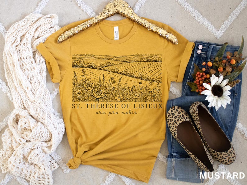 Pode incluir: Camiseta amarelo mostarda com um gr&aacute;fico de paisagem preto e o texto "ST. THERESE OF LISIEUX ora pro nobis." A camisa &eacute; combinada com jeans, sapatilhas com estampa de leopardo e um arranjo floral.
