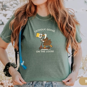 May include: Moss green t-shirt featuring a cartoon goose riding a scooter and holding a flag. The text on the shirt reads "Catholic Goose On The Loose". The goose is wearing a brown robe and the flag has a yellow and white design.