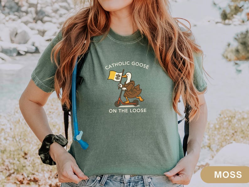 May include: Moss green t-shirt featuring a cartoon goose riding a scooter and holding a flag. The text on the shirt reads "Catholic Goose On The Loose". The goose is wearing a brown robe and the flag has a yellow and white design.