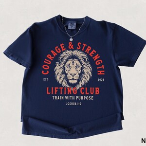 May include: Navy blue t-shirt with a lion's head graphic and the words "COURAGE & STRENGTH" in a circular design. The text "LIFTING CLUB" and "TRAIN WITH PURPOSE" are also displayed. The shirt includes the text "JOSHUA 1:9".