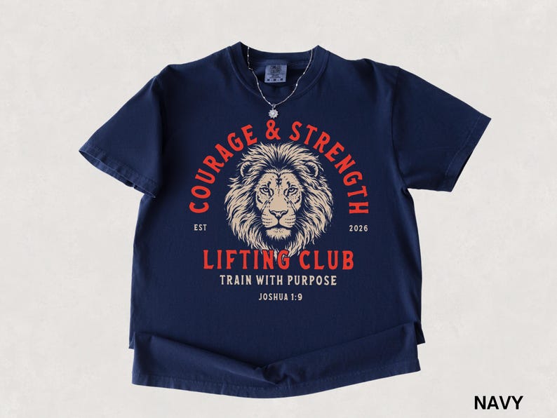 May include: Navy blue t-shirt with a lion's head graphic and the words "COURAGE & STRENGTH" in a circular design. The text "LIFTING CLUB" and "TRAIN WITH PURPOSE" are also displayed. The shirt includes the text "JOSHUA 1:9".