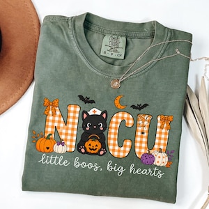 May include: An olive green t-shirt featuring the text NOCU in orange and white gingham letters. A black cat wearing a nurse cap holds a jack-o'-lantern. The design includes pumpkins, bats, a spider web, and the text little boos, big hearts.