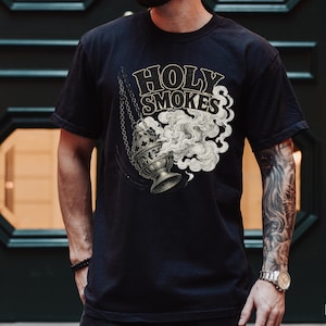 May include: Black t-shirt featuring a graphic of a censer with smoke and the words "Holy Smokes" in a vintage style. The design is in shades of gray and cream. The shirt is a classic crew neck.