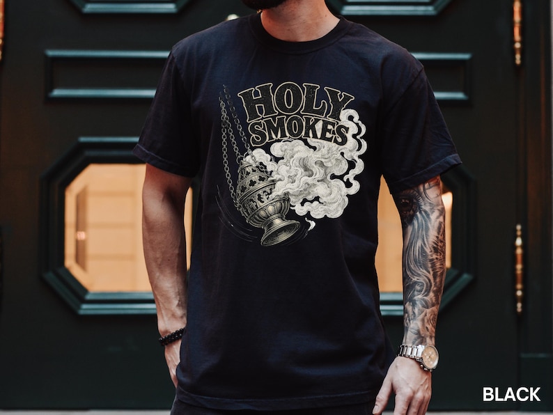 May include: Black t-shirt featuring a graphic of a censer with smoke and the words "Holy Smokes" in a vintage style. The design is in shades of gray and cream. The shirt is a classic crew neck.