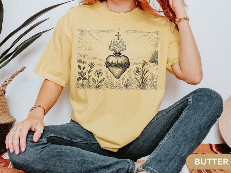K&ouml;nnte beinhalten: Hellgelbes T-Shirt mit einer Schwarz-Wei&szlig;-Grafik eines Herzens, Flammen und eines Kreuzes. Das Design ist vor einer Landschaft mit Blumen und H&uuml;geln platziert. Das Wort "BUTTER" ist unten rechts zu sehen.