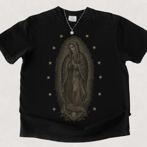 Puede incluir: Camiseta negra con una ilustraci&oacute;n en tonos sepia de la Virgen de Guadalupe, rodeada de estrellas. Se ve un collar plateado con un colgante. La camiseta tiene mangas cortas y cuello redondo.