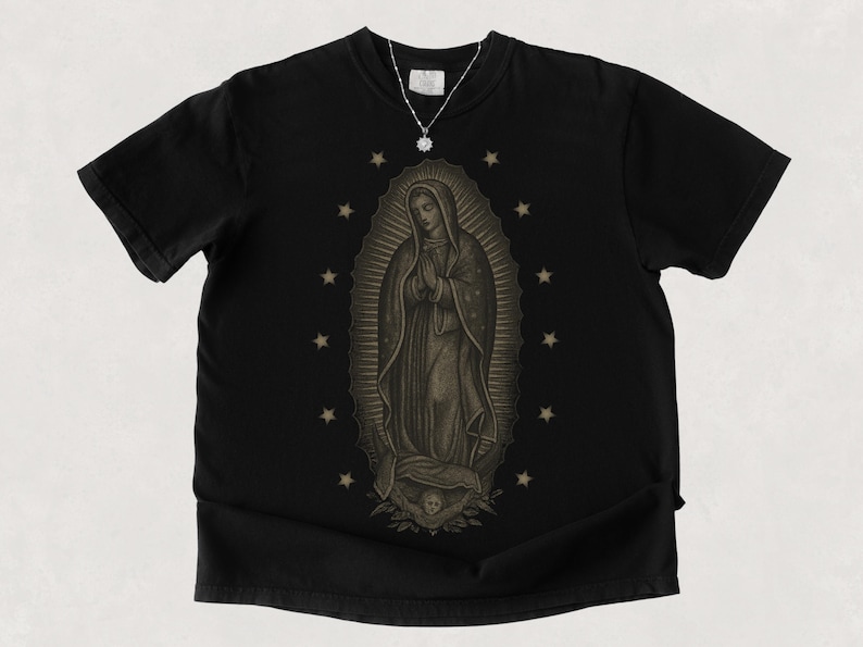 Puede incluir: Camiseta negra con una ilustraci&oacute;n en tonos sepia de la Virgen de Guadalupe, rodeada de estrellas. Se ve un collar plateado con un colgante. La camiseta tiene mangas cortas y cuello redondo.