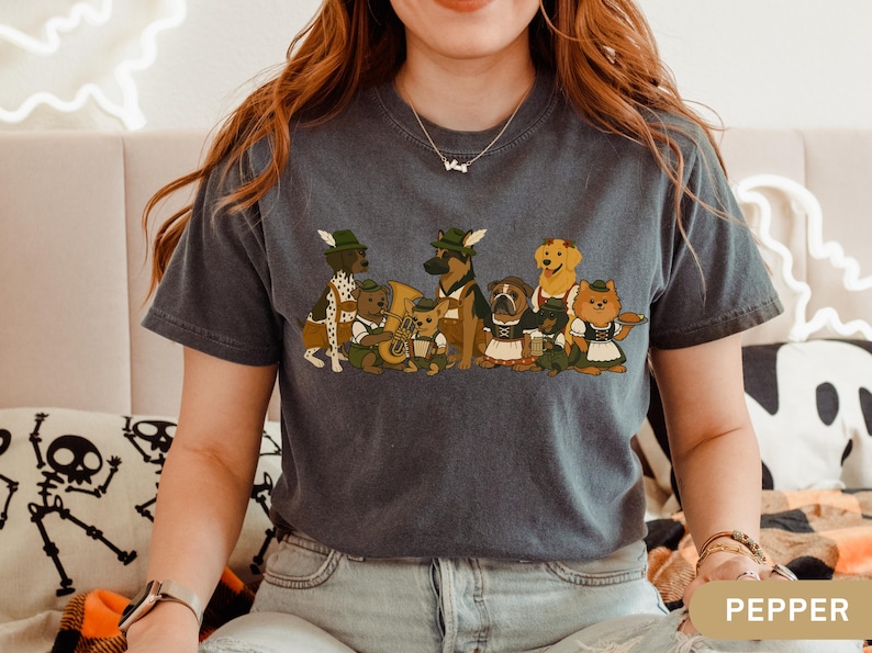 May include: Dark grey t-shirt featuring a cartoon illustration of various dog breeds dressed in traditional Bavarian attire. The dogs are depicted with hats, lederhosen, and instruments, creating a festive and whimsical design. The word "PEPPER" is printed on the bottom right.