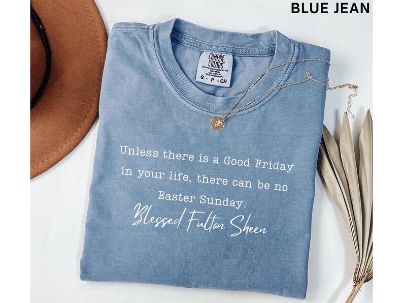 May include: A blue jean coloured t-shirt with the text "Unless there is a Good Friday in your life, there can be no Easter Sunday. Blessed Fulton Sheen." A gold necklace with a pendant is on the shirt.