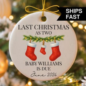 May include: A round ceramic Christmas ornament with a gold string for hanging. The ornament features the text "LAST CHRISTMAS AS TWO" and "BABY WILLIAMS IS DUE June 2026". It has a festive design with three red Christmas stockings.