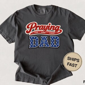 Puede incluir: Camiseta gris oscuro con las palabras "Praying DAD" en letras rojas, blancas y azules. La palabra "Praying" está en cursiva, y "DAD" está en letras de bloque con un patrón de estrellas. La camiseta tiene cuello redondo.