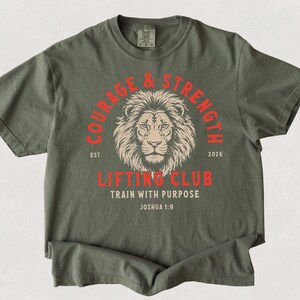 May include: Moss green t-shirt with a lion graphic and the words "COURAGE & STRENGTH" in a red arch. Below the lion is "LIFTING CLUB TRAIN WITH PURPOSE" in red. The text "EST 2026" is also present.