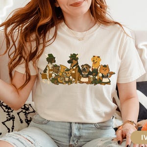 May include: Ivory-colored t-shirt featuring a cartoon illustration of various dog breeds dressed in traditional German attire. The dogs are depicted in a line, some holding instruments, and others wearing hats and aprons. The shirt is made of a soft material.