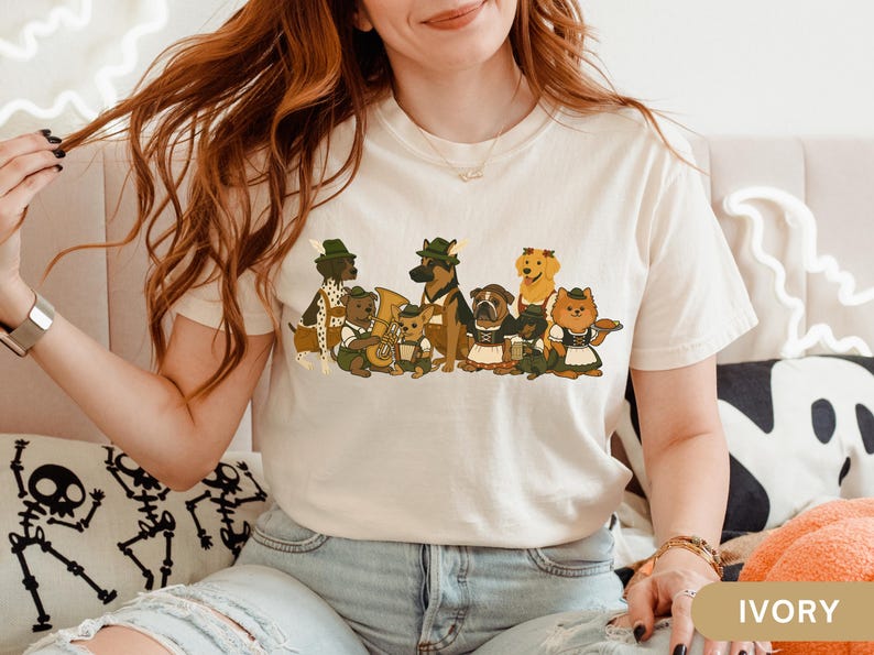 May include: Ivory-colored t-shirt featuring a cartoon illustration of various dog breeds dressed in traditional German attire. The dogs are depicted in a line, some holding instruments, and others wearing hats and aprons. The shirt is made of a soft material.