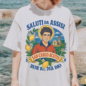 May include: White t-shirt with a vibrant illustration. The design includes the text "SALUTI DA ASSISI" and "SAN CARLO ACUTIS", a portrait, lemons, and a church. This casual t-shirt is suitable for everyday wear.