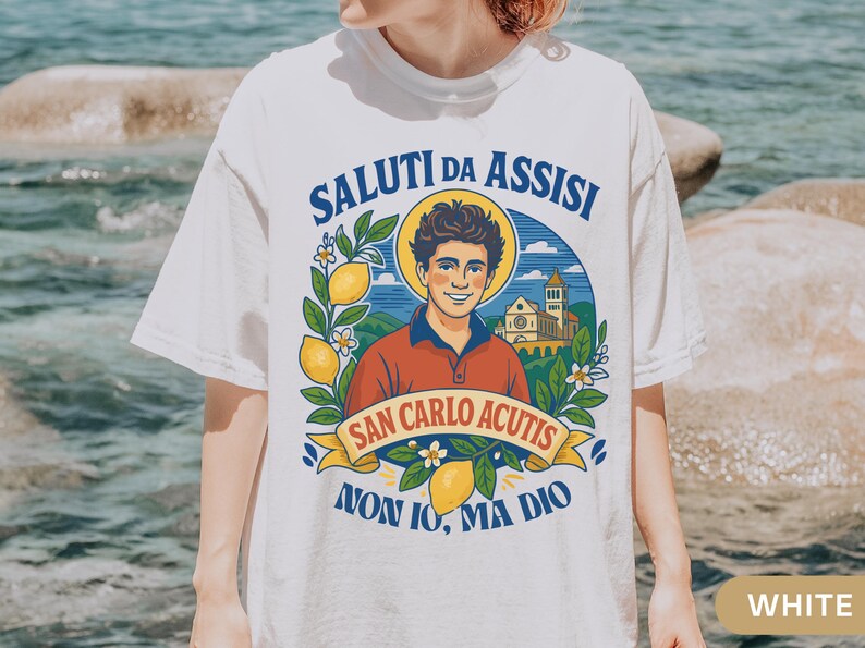 May include: White t-shirt with a vibrant illustration. The design includes the text "SALUTI DA ASSISI" and "SAN CARLO ACUTIS", a portrait, lemons, and a church. This casual t-shirt is suitable for everyday wear.