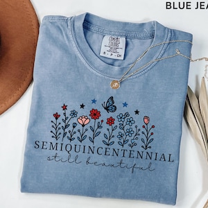May include: A light blue t-shirt with a floral design featuring red, pink, and blue flowers, a butterfly, and the text "SEMIQUINCENTENNIAL still beautiful." A gold necklace with a pendant is draped across the shirt.