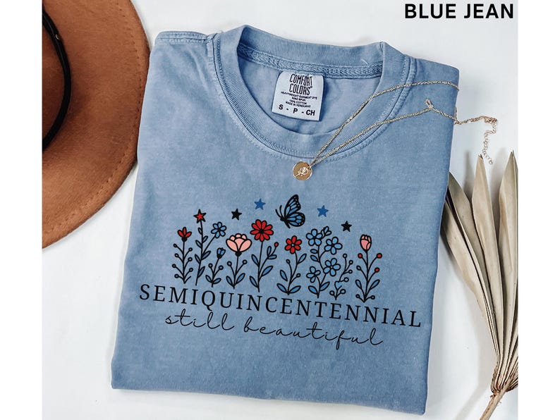 May include: A light blue t-shirt with a floral design featuring red, pink, and blue flowers, a butterfly, and the text "SEMIQUINCENTENNIAL still beautiful." A gold necklace with a pendant is draped across the shirt.