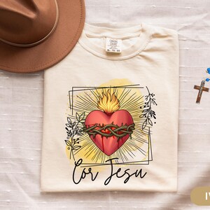 May include: Ivory-colored t-shirt featuring a graphic of a red heart with a crown of thorns, flames, and the text "Cor Jesu." A brown hat and a blue rosary with a cross are also visible.
