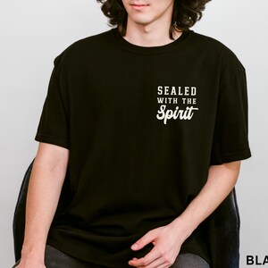 May include: Black t-shirt with the text "SEALED WITH THE Spirit" in white script. The shirt is a solid black color and has a crew neck. The text is centered on the chest.