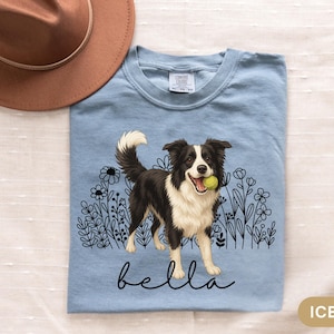 Könnte beinhalten: Hellblaues T-Shirt mit einer Illustration eines schwarz-weißen Border Collies, der einen Tennisball hält, umgeben von floralen Designs und dem Namen "Bella". Ein brauner Hut und ein Tennisball sind ebenfalls sichtbar. Das Shirt ist mit "ICE BLUE" beschriftet.