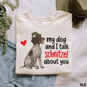 Può includere: Una t-shirt color naturale con la grafica di un cane bracco tedesco a pelo corto maculato che indossa un cappello alpino verde con un fiore. Il testo recita, my dog and I talk schnitzel about you, accompagnato da un piccolo cuore rosso.