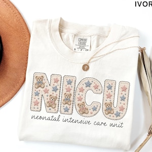 May include: Ivory-colored t-shirt with the letters "NICU" in a decorative font, featuring teddy bears and star accents. The words "neonatal intensive care unit" are printed below. A gold necklace and a brown hat are visible.