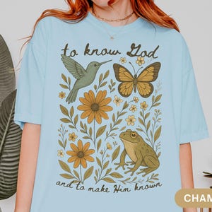 May include: Light blue t-shirt with a nature-themed graphic. The design includes a hummingbird, butterfly, frog, and flowers, with the text "to know God and to make Him known". The shirt is a relaxed fit.