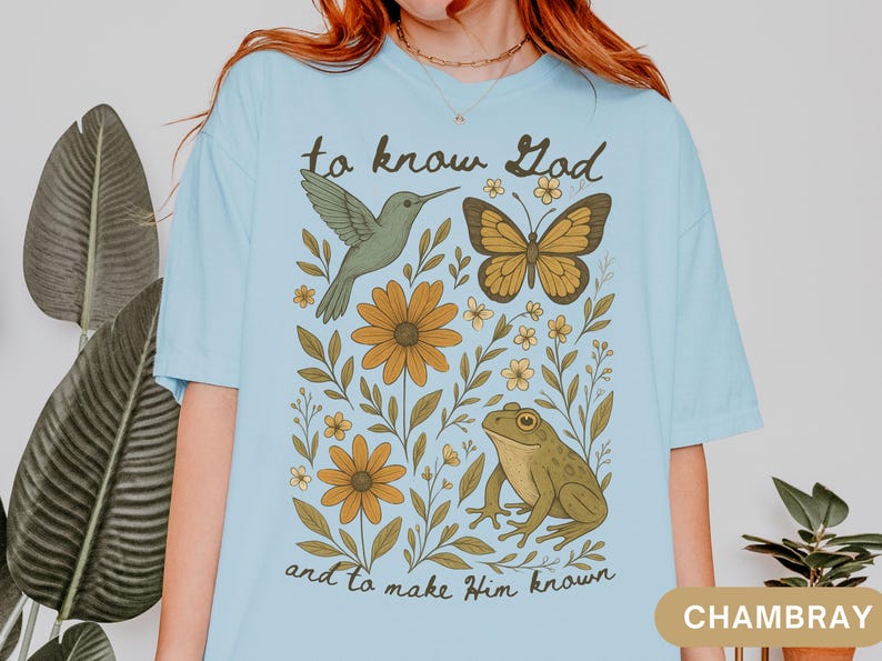 May include: Light blue t-shirt with a nature-themed graphic. The design includes a hummingbird, butterfly, frog, and flowers, with the text "to know God and to make Him known". The shirt is a relaxed fit.