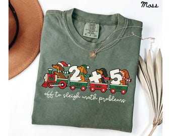 May include: A moss green t-shirt featuring a graphic of dachshunds riding a Christmas train. The dogs wear winter hats and scarves. The train cars carry the numbers 2, plus, and 5. The text reads, off to sleigh math problems.