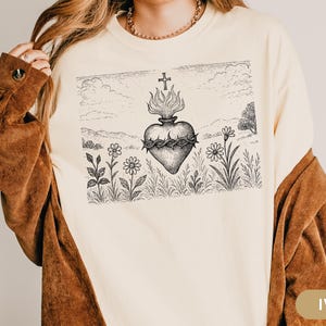K&ouml;nnte beinhalten: Elfenbeinfarbenes Langarmshirt mit einer detaillierten Schwarz-Wei&szlig;-Illustration eines Herzens mit Dornenkrone, Flammen und einem Kreuz. Das Design ist vor einer Landschaft mit Blumen und einem Kreuz platziert.