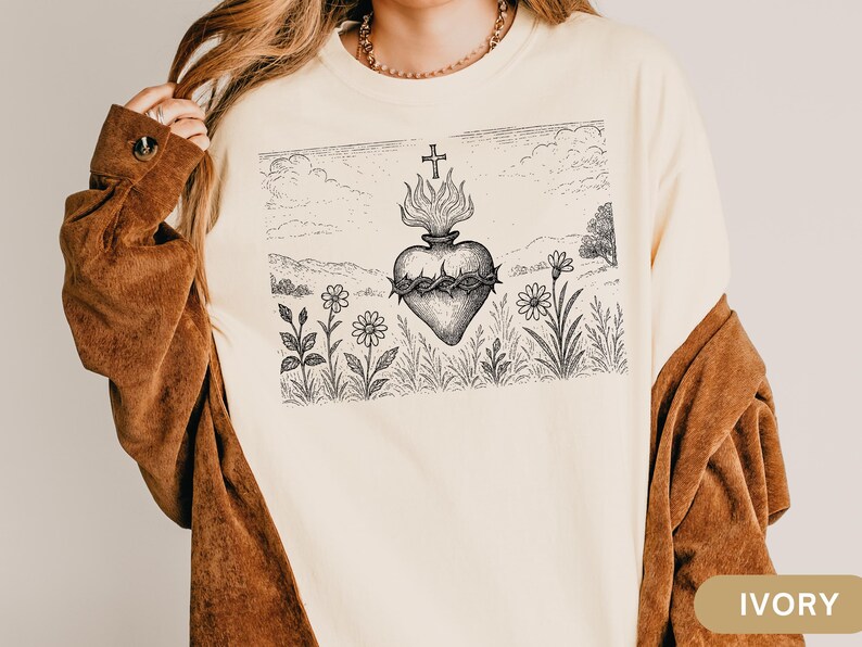 K&ouml;nnte beinhalten: Elfenbeinfarbenes Langarmshirt mit einer detaillierten Schwarz-Wei&szlig;-Illustration eines Herzens mit Dornenkrone, Flammen und einem Kreuz. Das Design ist vor einer Landschaft mit Blumen und einem Kreuz platziert.