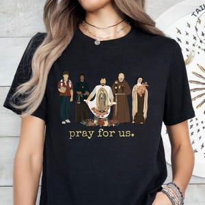 May include: Black t-shirt with a graphic of five figures in religious attire, including a woman holding a baby. The text "pray for us." is printed below the graphic.