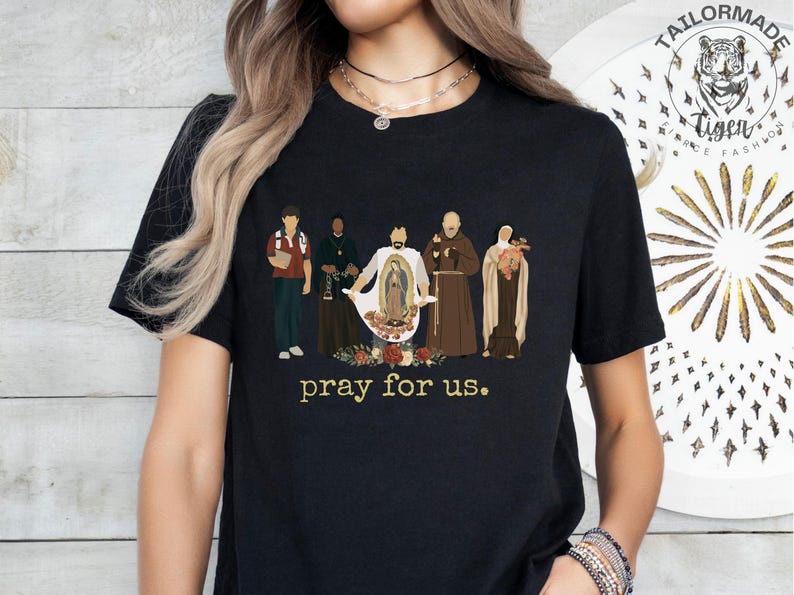 May include: Black t-shirt with a graphic of five figures in religious attire, including a woman holding a baby. The text "pray for us." is printed below the graphic.