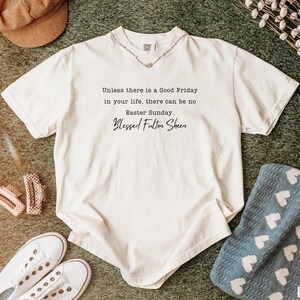 May include: Cream-coloured t-shirt featuring the quote "Unless there is a Good Friday in your life, there can be no Easter Sunday. Blessed Fulton Sheen." The shirt is styled with white trainers, hair clips, and a brown hat.