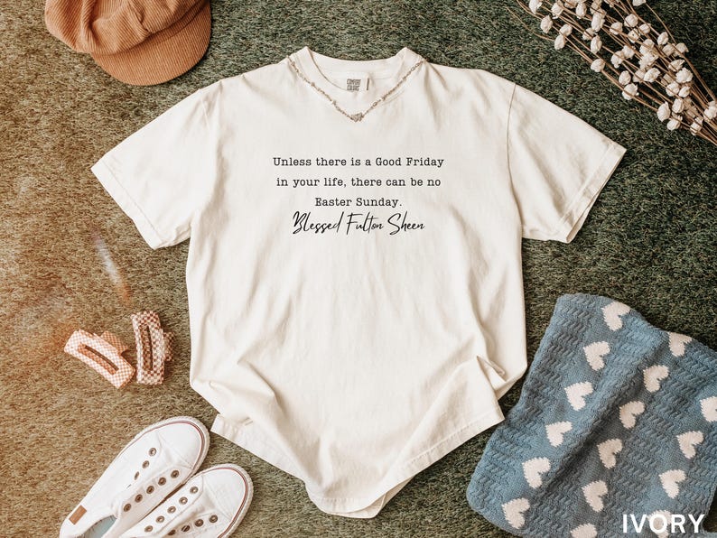 May include: Cream-coloured t-shirt featuring the quote "Unless there is a Good Friday in your life, there can be no Easter Sunday. Blessed Fulton Sheen." The shirt is styled with white trainers, hair clips, and a brown hat.