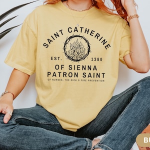 Puede incluir: Una camiseta amarilla con texto negro que dice "Saint Catherine Est. 1380 Of Sienna Patron Saint Of Nurses, The Sick & Fire Prevention". El texto está rodeado por una corona con una llama en el centro.