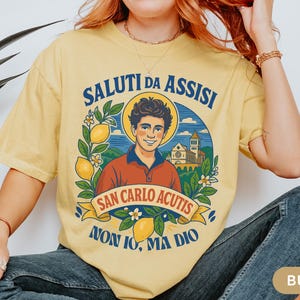 May include: Pale yellow t-shirt with a graphic design including the text "SALUTI DA ASSISI" and "SAN CARLO ACUTIS". The design features a man, lemons, and a church. The t-shirt is worn by a person.