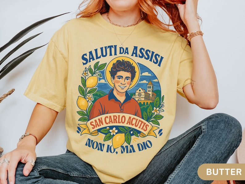 May include: Pale yellow t-shirt with a graphic design including the text "SALUTI DA ASSISI" and "SAN CARLO ACUTIS". The design features a man, lemons, and a church. The t-shirt is worn by a person.